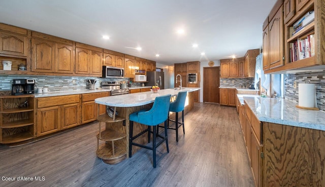 kitchen with brown cabinets, open shelves, stainless steel appliances, dark wood-type flooring, and a kitchen island with sink