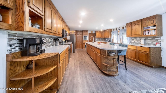 kitchen with light countertops, appliances with stainless steel finishes, open shelves, brown cabinetry, and an island with sink