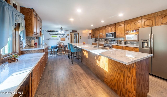 kitchen with brown cabinets, dark wood-style flooring, a kitchen island with sink, stainless steel appliances, and a sink