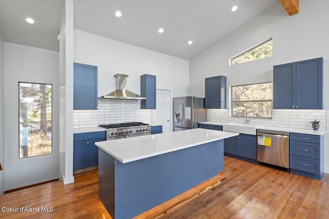 kitchen with stainless steel appliances, a kitchen island, light countertops, wall chimney range hood, and blue cabinetry