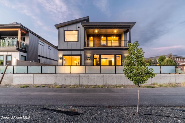 back of house at dusk featuring a balcony, a fenced front yard, and board and batten siding