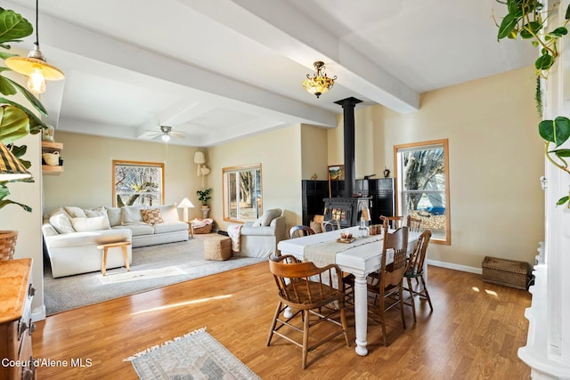 dining area with ceiling fan, wood finished floors, baseboards, beamed ceiling, and a wood stove