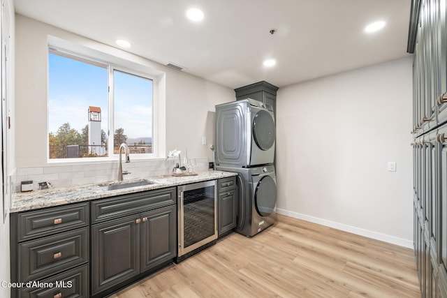 clothes washing area with laundry area, stacked washer / dryer, wine cooler, light wood-type flooring, and a sink