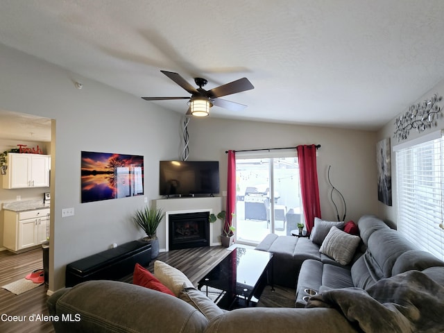 living area featuring vaulted ceiling, a fireplace, a textured ceiling, a ceiling fan, and dark wood-style flooring