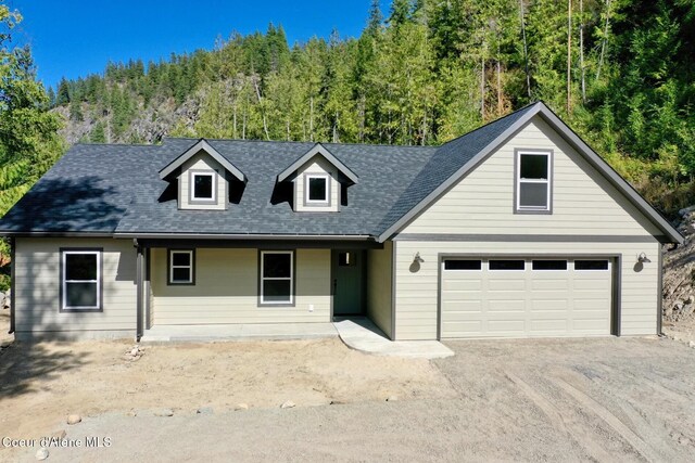 view of front facade with a garage, driveway, roof with shingles, and a view of trees