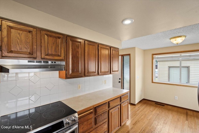 kitchen featuring under cabinet range hood, electric stove, decorative backsplash, light wood finished floors, and brown cabinetry