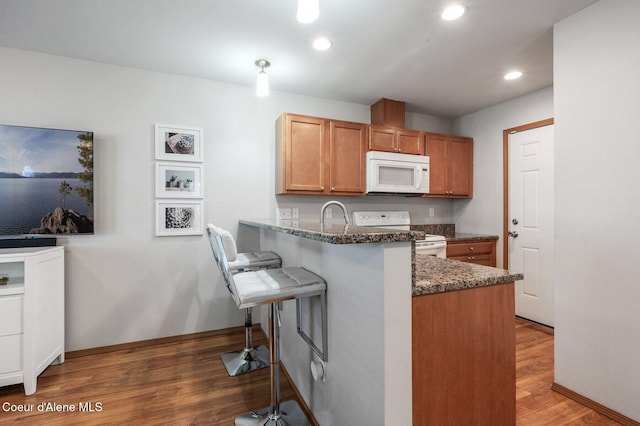 kitchen with dark stone counters, a kitchen bar, a peninsula, wood finished floors, and white appliances