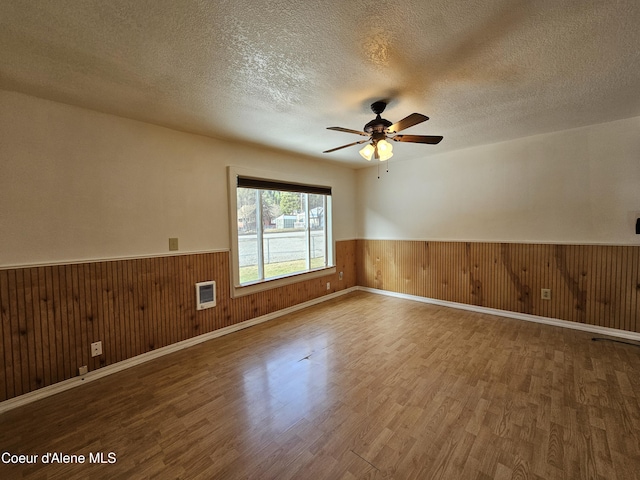 empty room with a ceiling fan, wainscoting, wood walls, a textured ceiling, and wood finished floors