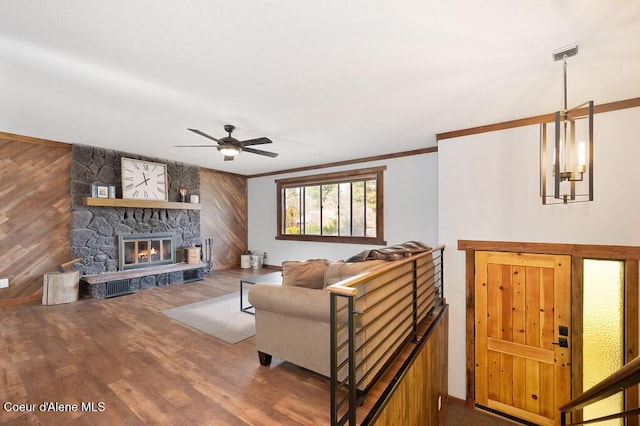 living room featuring ornamental molding, ceiling fan with notable chandelier, wood finished floors, a stone fireplace, and wood walls