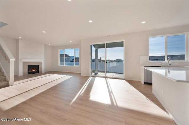 unfurnished living room with recessed lighting, light wood-style flooring, a sink, a warm lit fireplace, and stairs