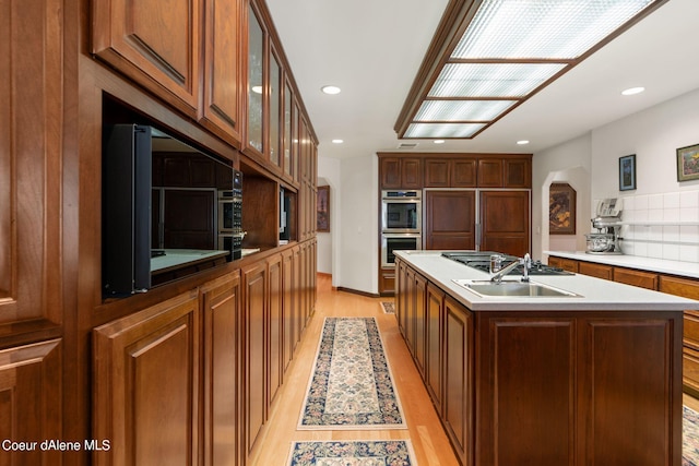 kitchen with arched walkways, glass insert cabinets, stainless steel double oven, a kitchen island with sink, and light wood-type flooring