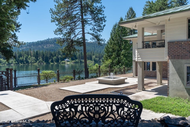 view of patio with a wooded view, an outdoor fire pit, a water view, and fence