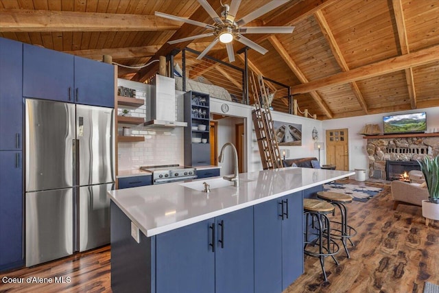 kitchen featuring wall chimney range hood, stainless steel appliances, a stone fireplace, blue cabinetry, and a sink