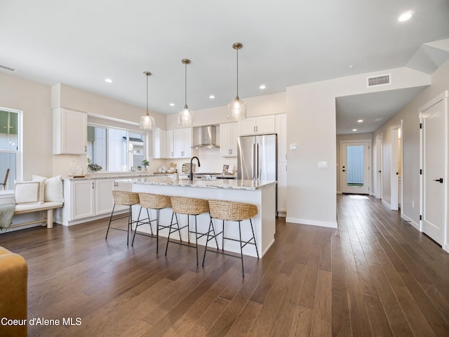 kitchen featuring a kitchen island with sink, dark wood-style flooring, stainless steel refrigerator, white cabinets, and wall chimney exhaust hood