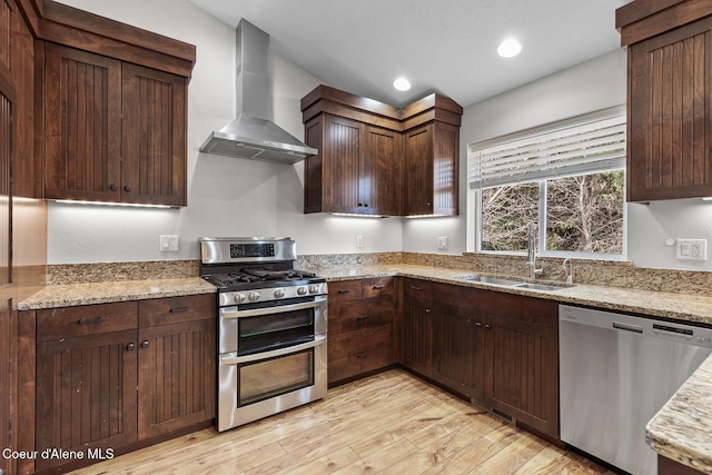 kitchen with appliances with stainless steel finishes, a sink, wall chimney range hood, light stone countertops, and light wood-type flooring