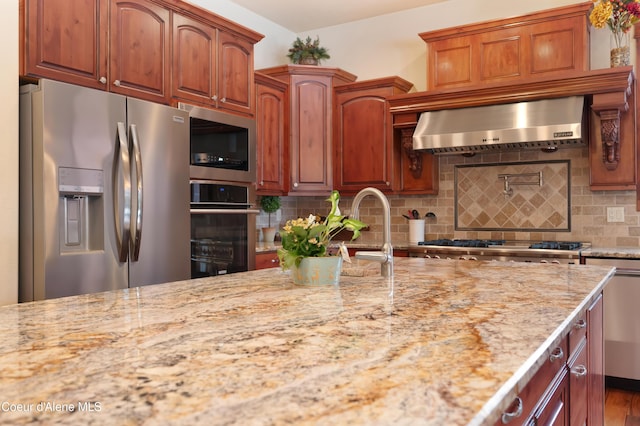 kitchen with decorative backsplash, stainless steel appliances, wall chimney exhaust hood, and light stone countertops