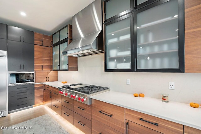 kitchen featuring light countertops, wall chimney exhaust hood, glass insert cabinets, and built in appliances