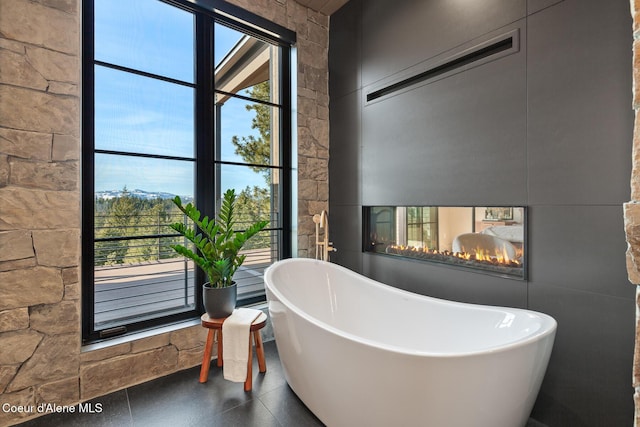 bathroom with a multi sided fireplace, a soaking tub, and tile walls