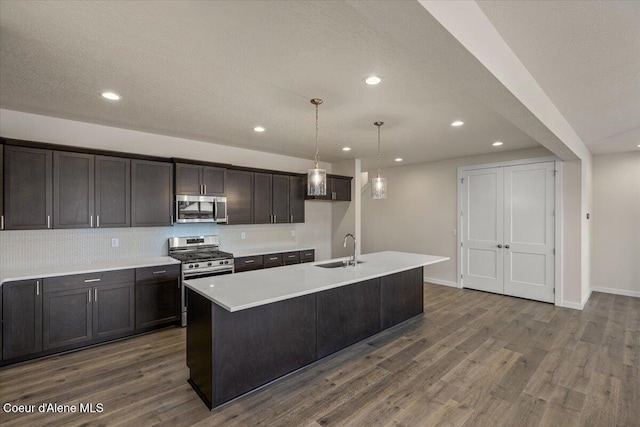 kitchen with a center island with sink, dark wood-style flooring, a sink, light countertops, and appliances with stainless steel finishes