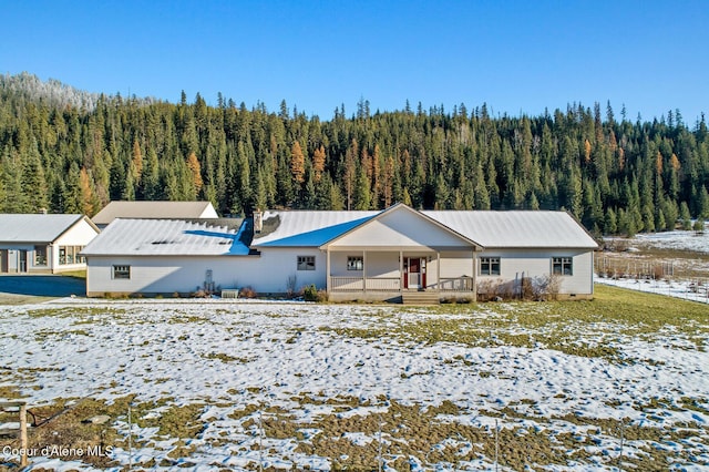 snow covered house featuring covered porch, a forest view, and metal roof