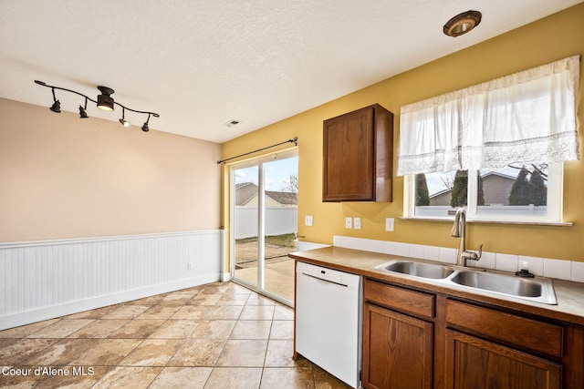 kitchen with a wainscoted wall, brown cabinets, a sink, a textured ceiling, and white dishwasher