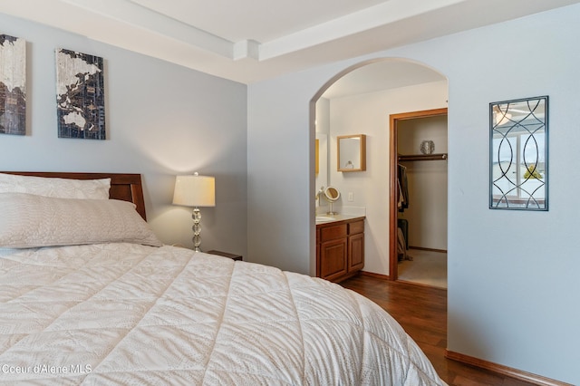 bedroom with a spacious closet, a closet, ensuite bathroom, arched walkways, and dark wood-style flooring