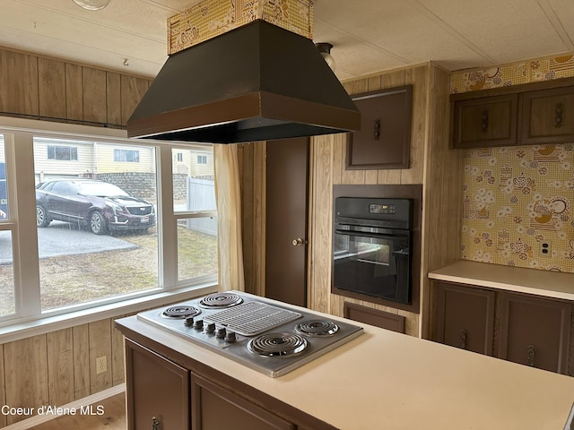 kitchen featuring oven, stainless steel electric cooktop, wooden walls, island range hood, and light countertops