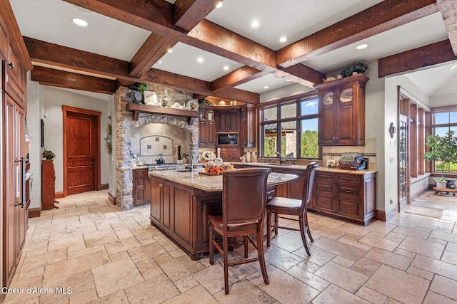 kitchen with beam ceiling, baseboards, stone tile floors, and an island with sink