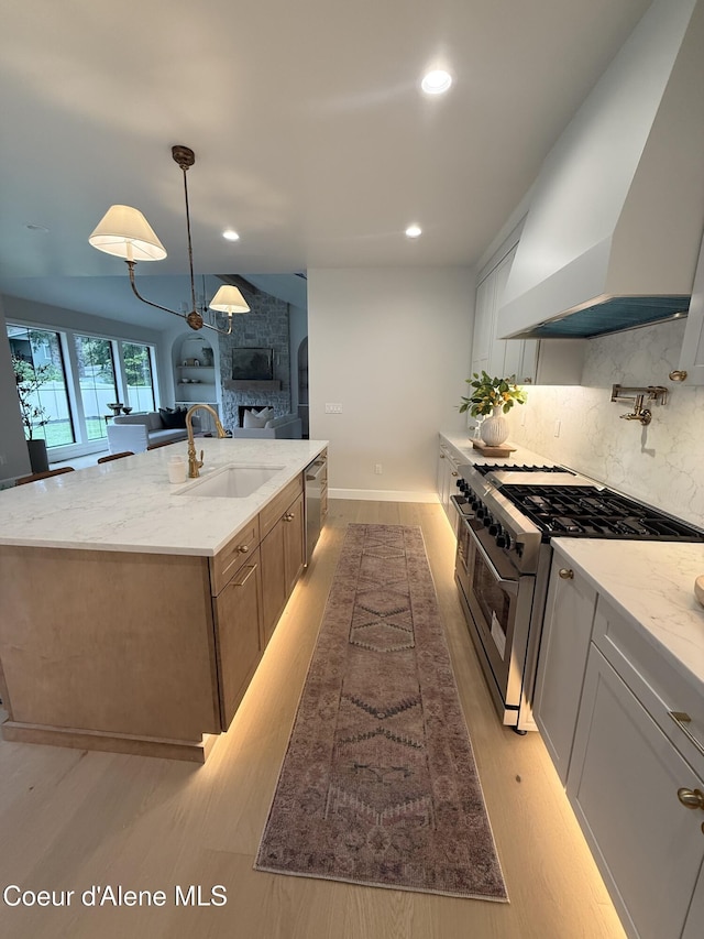 kitchen with appliances with stainless steel finishes, a sink, wall chimney exhaust hood, a fireplace, and a center island with sink