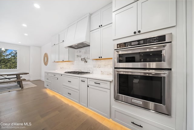 kitchen with light countertops, stainless steel double oven, gas cooktop, custom range hood, and backsplash