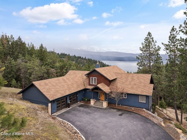 chalet / cabin featuring aphalt driveway, a water view, an attached garage, a chimney, and a shingled roof