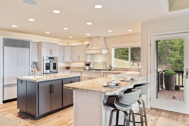 kitchen featuring gray cabinets, a sink, a center island with sink, custom exhaust hood, and stainless steel appliances