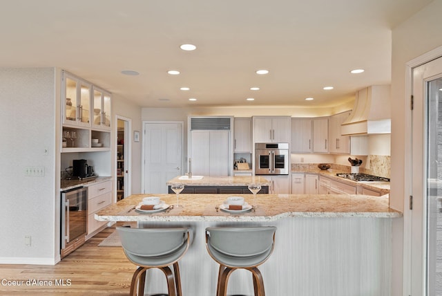 kitchen with light wood finished floors, wine cooler, stainless steel appliances, custom range hood, and a kitchen breakfast bar