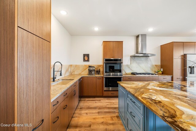 kitchen with decorative backsplash, light stone countertops, appliances with stainless steel finishes, a sink, and wall chimney range hood