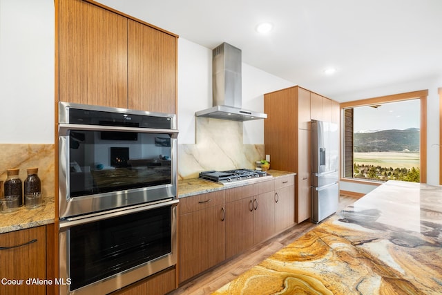 kitchen with decorative backsplash, light stone counters, light wood-style flooring, appliances with stainless steel finishes, and wall chimney exhaust hood