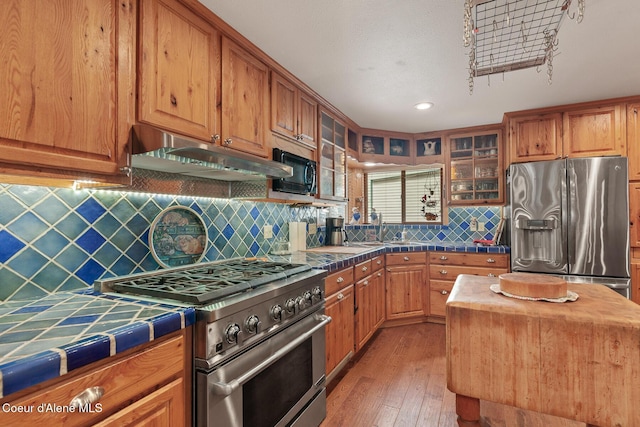kitchen with appliances with stainless steel finishes, glass insert cabinets, tile counters, under cabinet range hood, and light wood-style flooring
