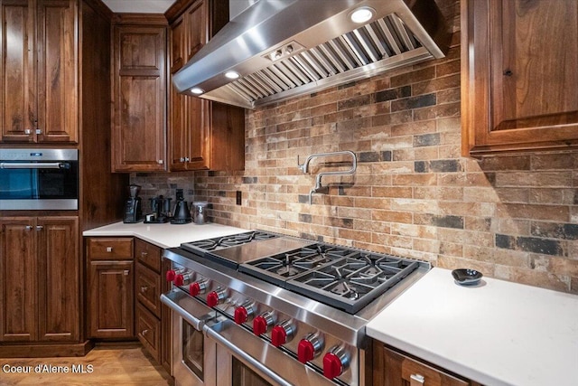 kitchen featuring wall chimney range hood, light countertops, light wood finished floors, stainless steel appliances, and decorative backsplash