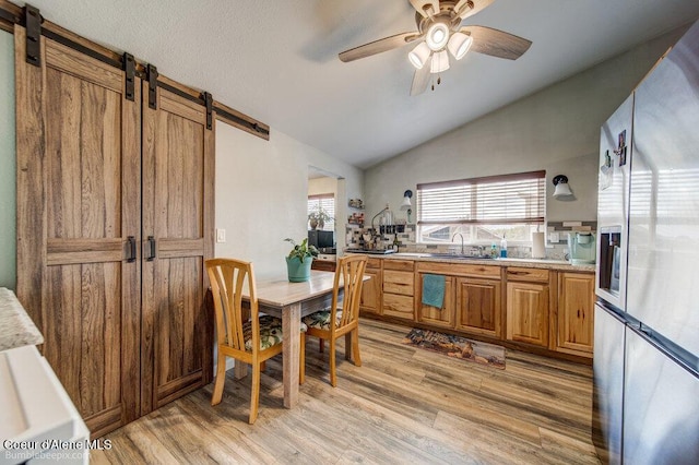 kitchen featuring ceiling fan, fridge with ice dispenser, light wood-style flooring, vaulted ceiling, and a barn door