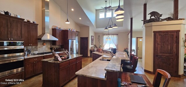 kitchen featuring high vaulted ceiling, wall chimney range hood, a spacious island, stainless steel appliances, and a sink