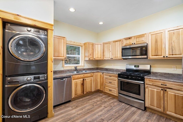 kitchen featuring stacked washer and clothes dryer, stainless steel appliances, dark countertops, wood finished floors, and a sink