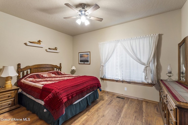 bedroom with ceiling fan, a textured ceiling, baseboards, wood finished floors, and visible vents