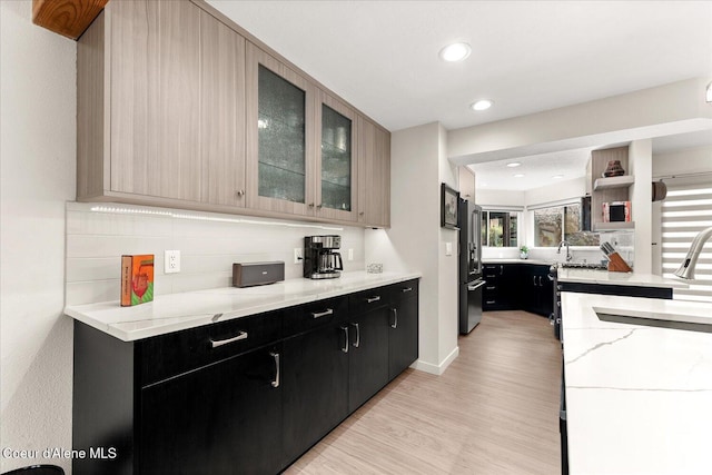 kitchen featuring light stone counters, light wood finished floors, dark cabinetry, recessed lighting, and glass insert cabinets