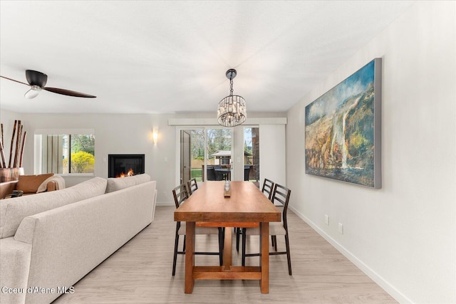 dining area with a glass covered fireplace, baseboards, ceiling fan with notable chandelier, and light wood-style flooring
