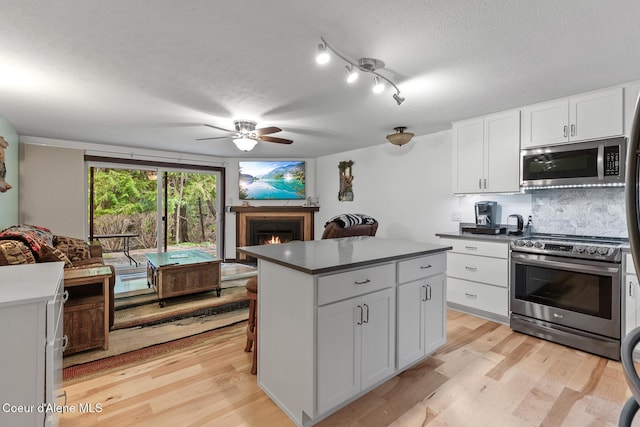 kitchen with a textured ceiling, light wood-style floors, stainless steel appliances, a warm lit fireplace, and a ceiling fan