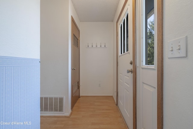 hallway with a textured ceiling, visible vents, light wood-type flooring, and a wainscoted wall