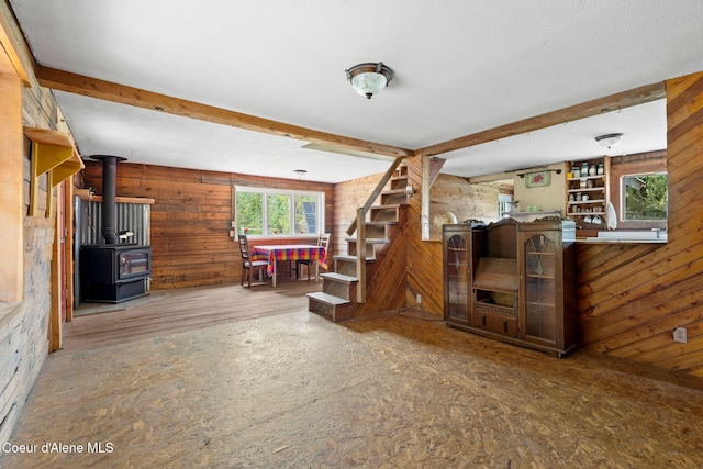 unfurnished living room featuring beamed ceiling, a wood stove, wood walls, and stairs