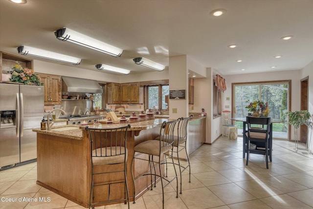 2885 S Folsom Ridge Rd, Coeur Dalene, ID 83814 - kitchen with stainless steel refrigerator with ice dispenser, wall chimney exhaust hood, brown cabinets, light stone countertops, and plenty of natural light