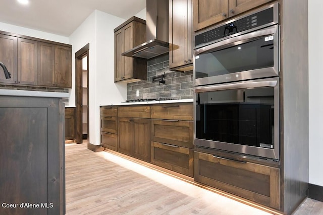 kitchen with wall chimney range hood, decorative backsplash, stainless steel double oven, light countertops, and light wood-style floors