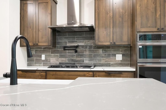 kitchen featuring decorative backsplash, double oven, wall chimney range hood, light countertops, and gas stovetop