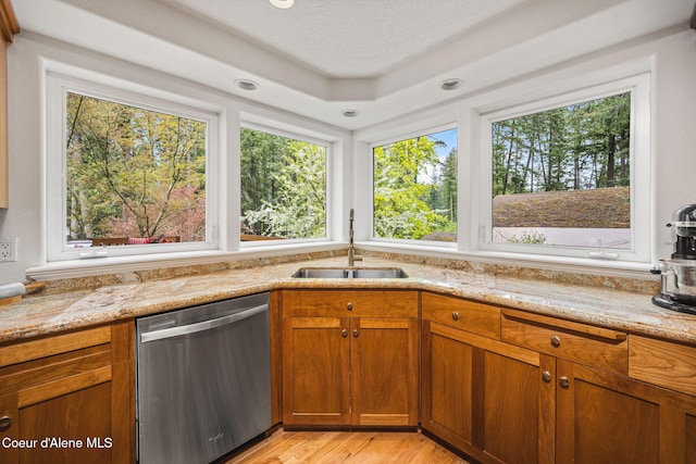 kitchen with brown cabinetry, light stone countertops, light wood-type flooring, a sink, and dishwasher